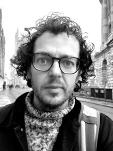 A close-up, black and white photo of me, Joshua, a white man with curly hair in his late thirties. I'm wearing a speckled woolen turtle-neck and black jacket, the strap of a rucksack hanging off one shoulder. I'm staring at the camera through my large black glasses, looking slightly surprised (even though I'm fairly sure this photo is a selfie).